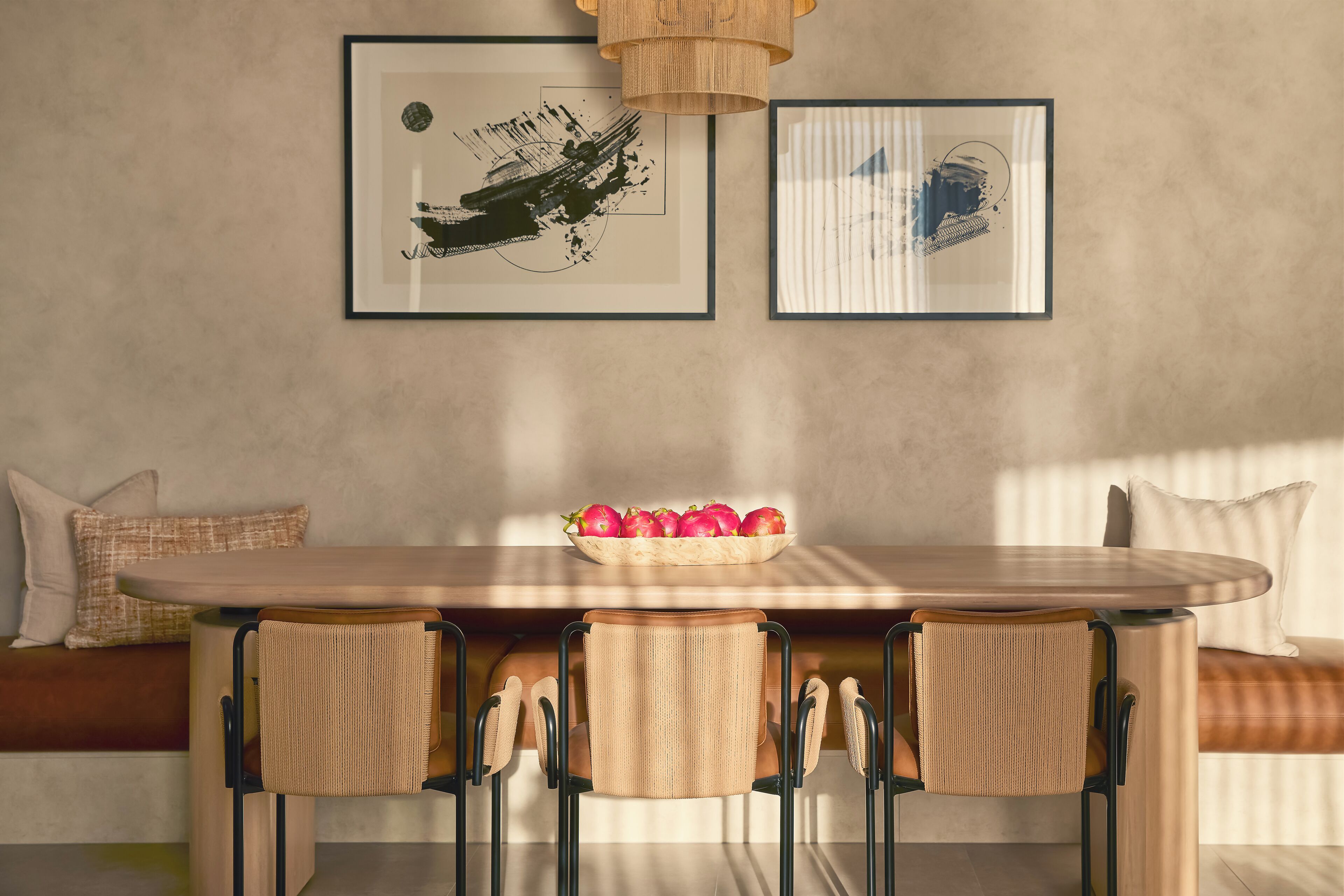 Un comedor con una mesa de madera, tres sillas tejidas, un cuenco de manzanas y dos cuadros en la pared.