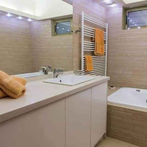 A modern bathroom with a white sink, a mirror, and a bathtub. There is a towel rack with towels and a window above the sink. - Panorama Luxury Villa in Sitia, Crete (Sitia)