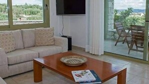 A living room with a sofa, a coffee table, a flat-screen TV, and a sliding glass door leading to an outdoor area with a table and chairs. - Panorama Luxury Villa in Sitia, Crete (Sitia)