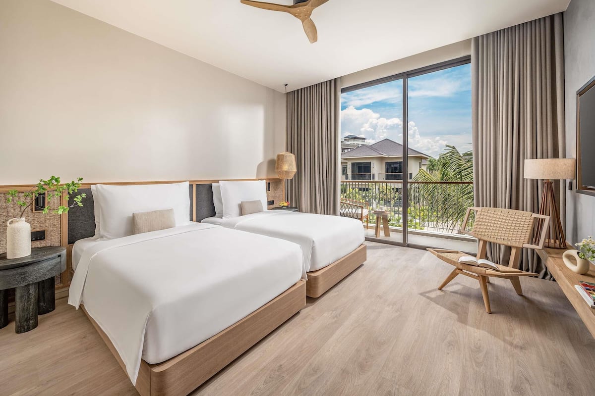 A modern hotel room with two beds, a ceiling fan, a balcony with a view, and a small table with a lamp.