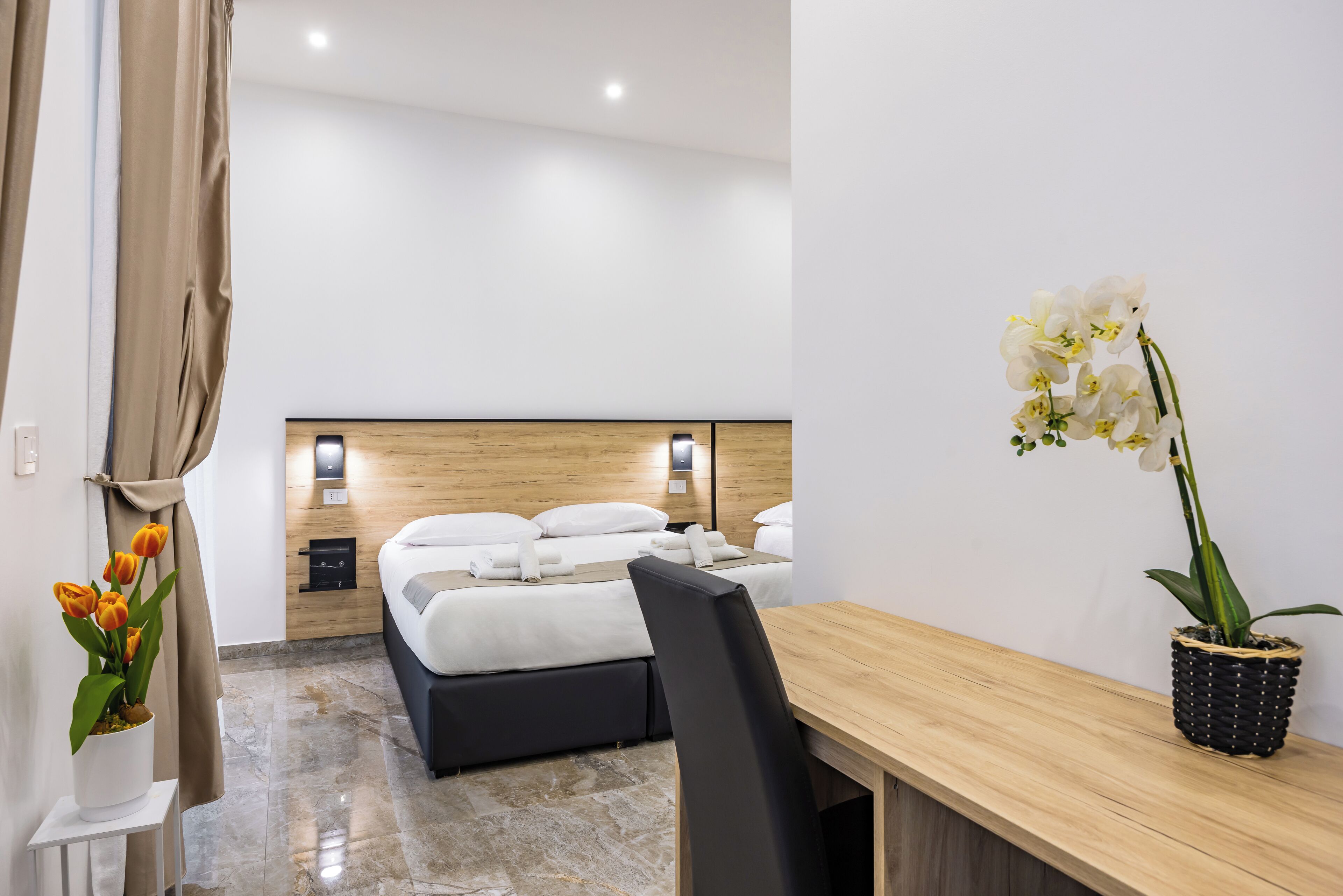 A hotel room with a bed, a desk, a chair, and a potted orchid.