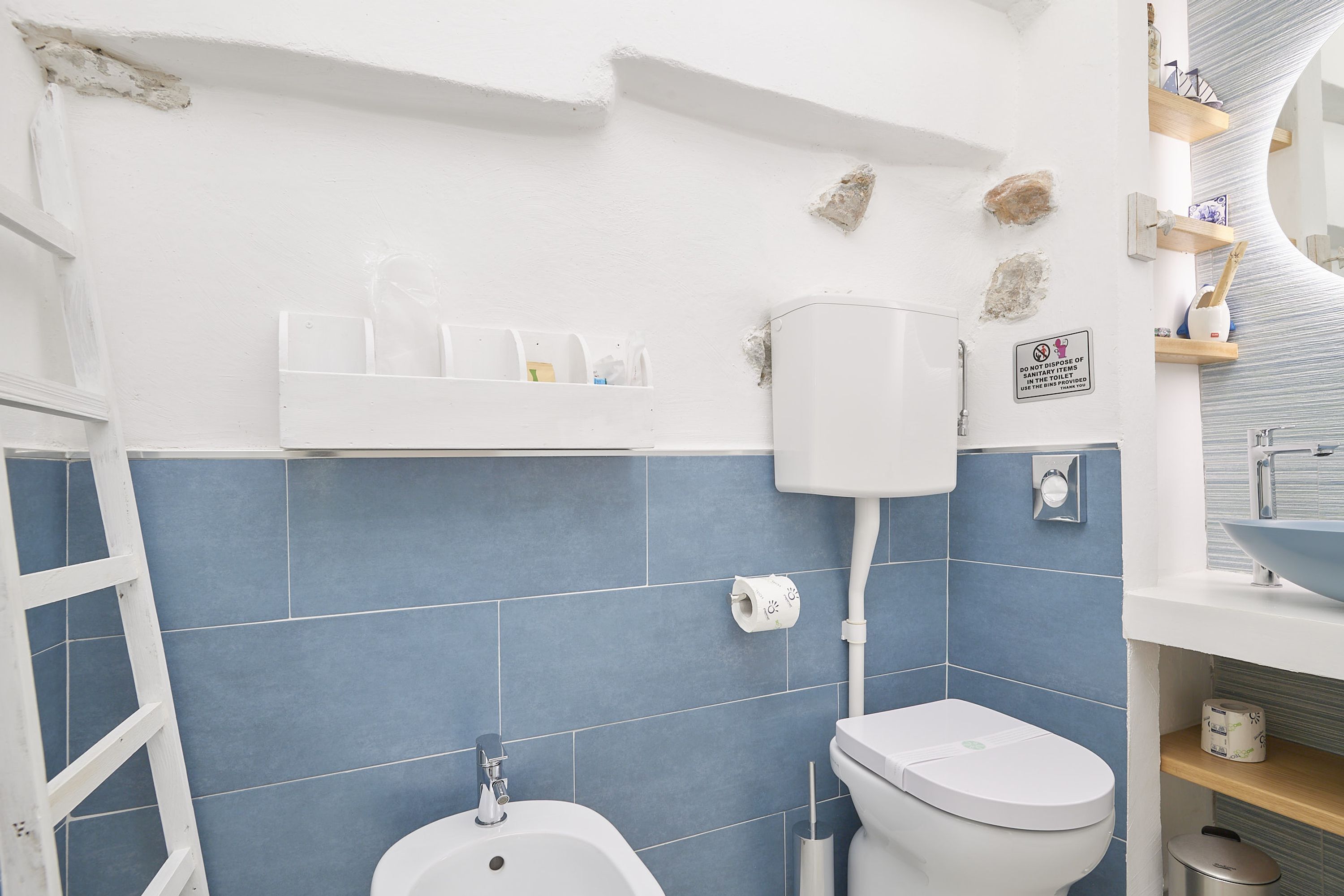 Une salle de bain équipée de toilettes, d’un bidet et d’un lavabo. On y trouve des étagères contenant des articles de toilette, ainsi qu’un panneau interdisant l’utilisation d’objets tranchants.