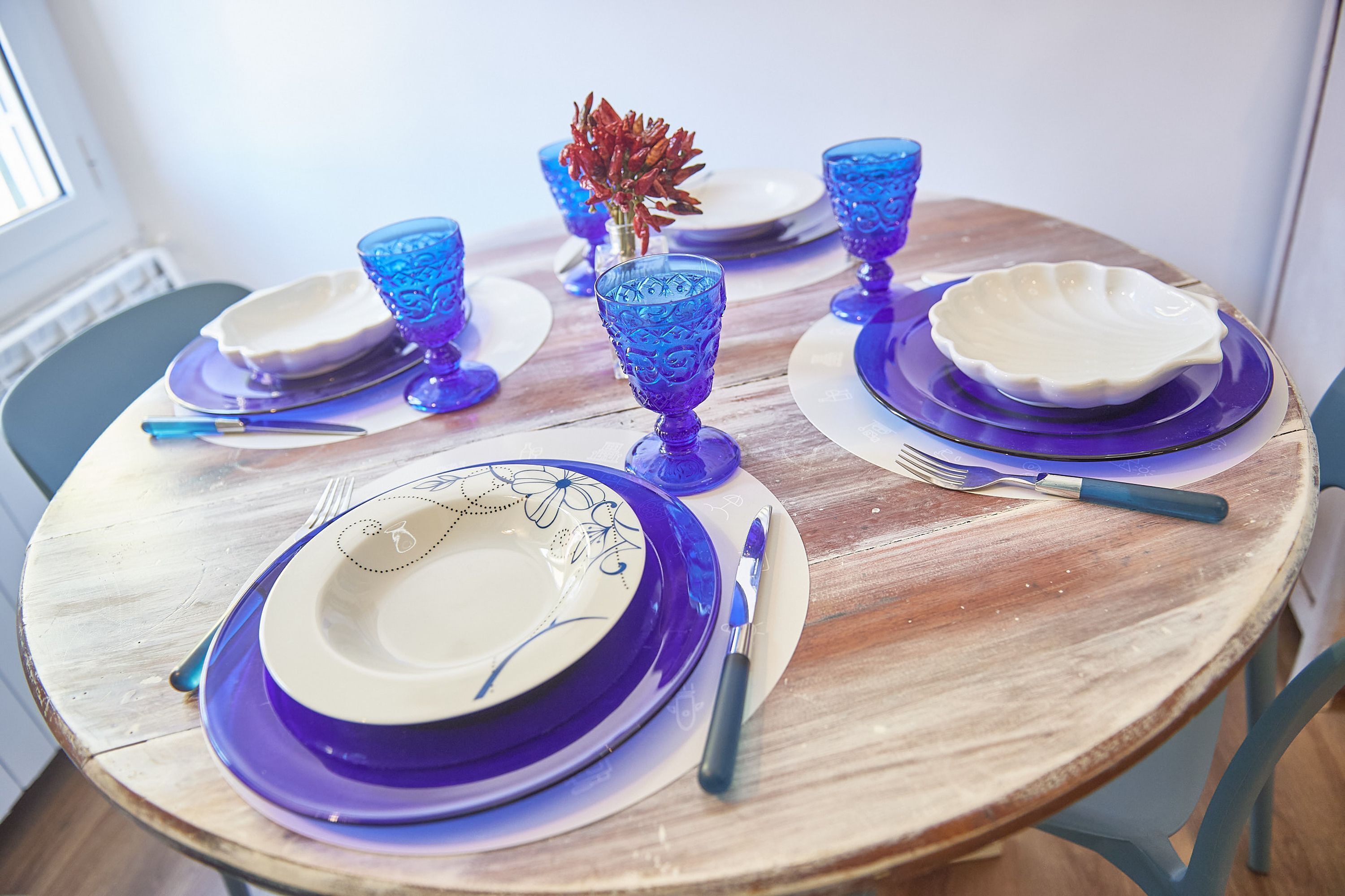 A round wooden table set with blue glassware, white plates, and a decorative red plant.