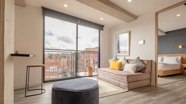 A modern hotel room with a large window, a sofa with cushions, a round ottoman, and a bed. - Chic 1 BR in Chapinero (Bogotá)