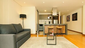 A modern living room with a grey sofa, a small table, and a kitchen in the background. - Cozy 2 Br apt in Candelaria (Bogotá)