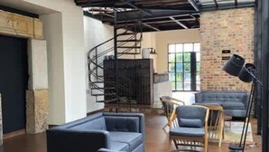 A spacious room with a high ceiling, exposed beams, a brick wall, and a large window. There are several seating areas with sofas and chairs, and a spiral staircase is visible in the background. - Stylish 2 Masters w Patio View (Bogotá)