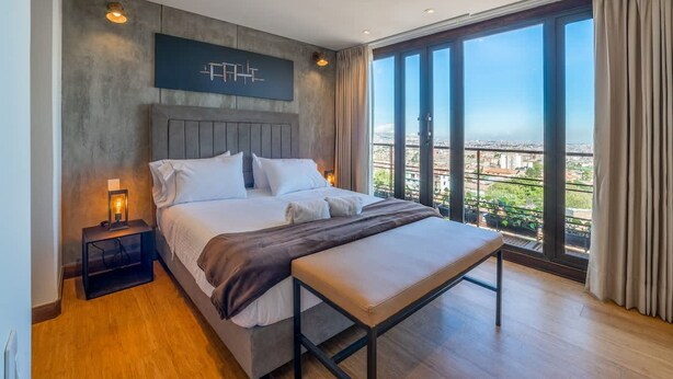 A modern bedroom with a large bed, a bench, a nightstand, and a city view through large windows. - Stylish 2 Masters w Patio View (Bogotá)
