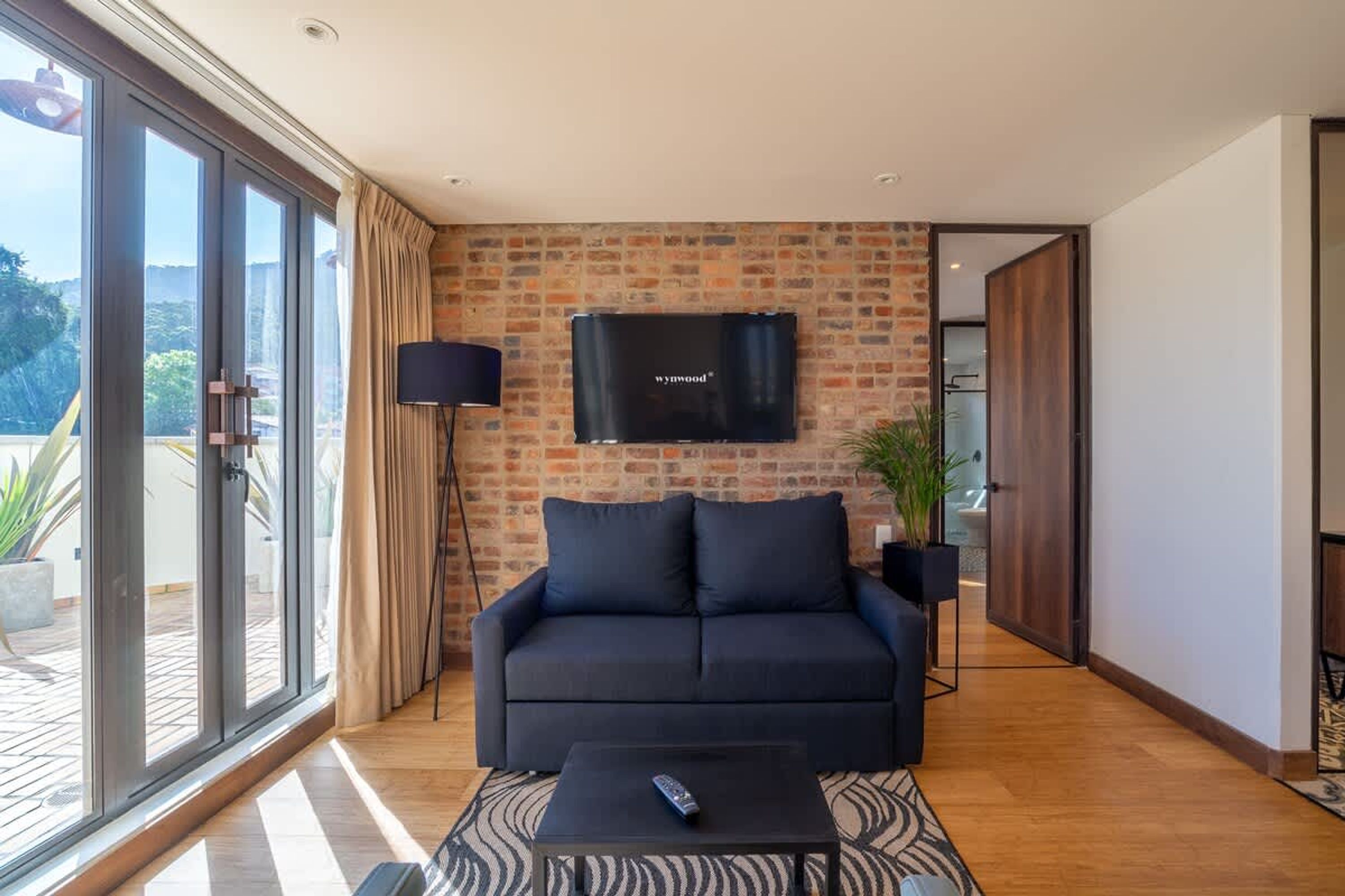 Una sala de estar moderna con un sofá, un televisor instalado en una pared de ladrillo, un control remoto sobre una mesa de centro y un cerramiento de vidrio corredero que da a un espacio exterior.