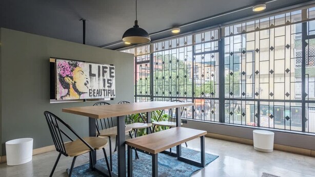 Ein Essbereich mit einem Holztisch, schwarzen Stühlen und einer Bank. Ein großes Fenster mit Metallgitterfenstern bietet Blick auf Gebäude. An der Wand hängt ein Bild mit dem Schriftzug "LIFE IS BEAUTIFUL".