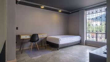 Quarto pequeno e moderno com uma cama, uma mesa e uma cadeira. Há uma janela com vista para edifícios e colinas.