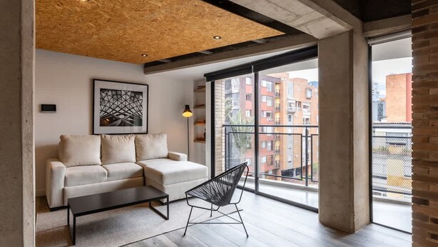 A modern living room with a sofa, a coffee table, and a chair. There is a large window with a view of buildings outside. - Stylish 1 BR in Chapinero (Bogotá)