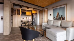 Ein modernes Wohnzimmer mit einem grauen Sofa, einem schwarzen Sessel und einem Holztisch. Im Hintergrund ist eine Küche mit Mikrowelle und Kühlschrank zu sehen.