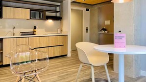 Una cocina moderna con armarios de madera, un microondas y un lavamanos. Hay una silla transparente y una silla blanca cerca de una mesita con un cartel de bienvenida.