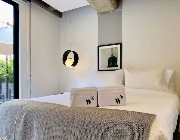 A modern hotel room with a large bed, a hanging light fixture, a framed picture on the wall, and a view of greenery outside.