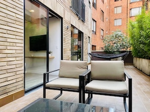 A modern outdoor seating area with two chairs and a glass table, adjacent to a building with large windows and a flat-screen TV.