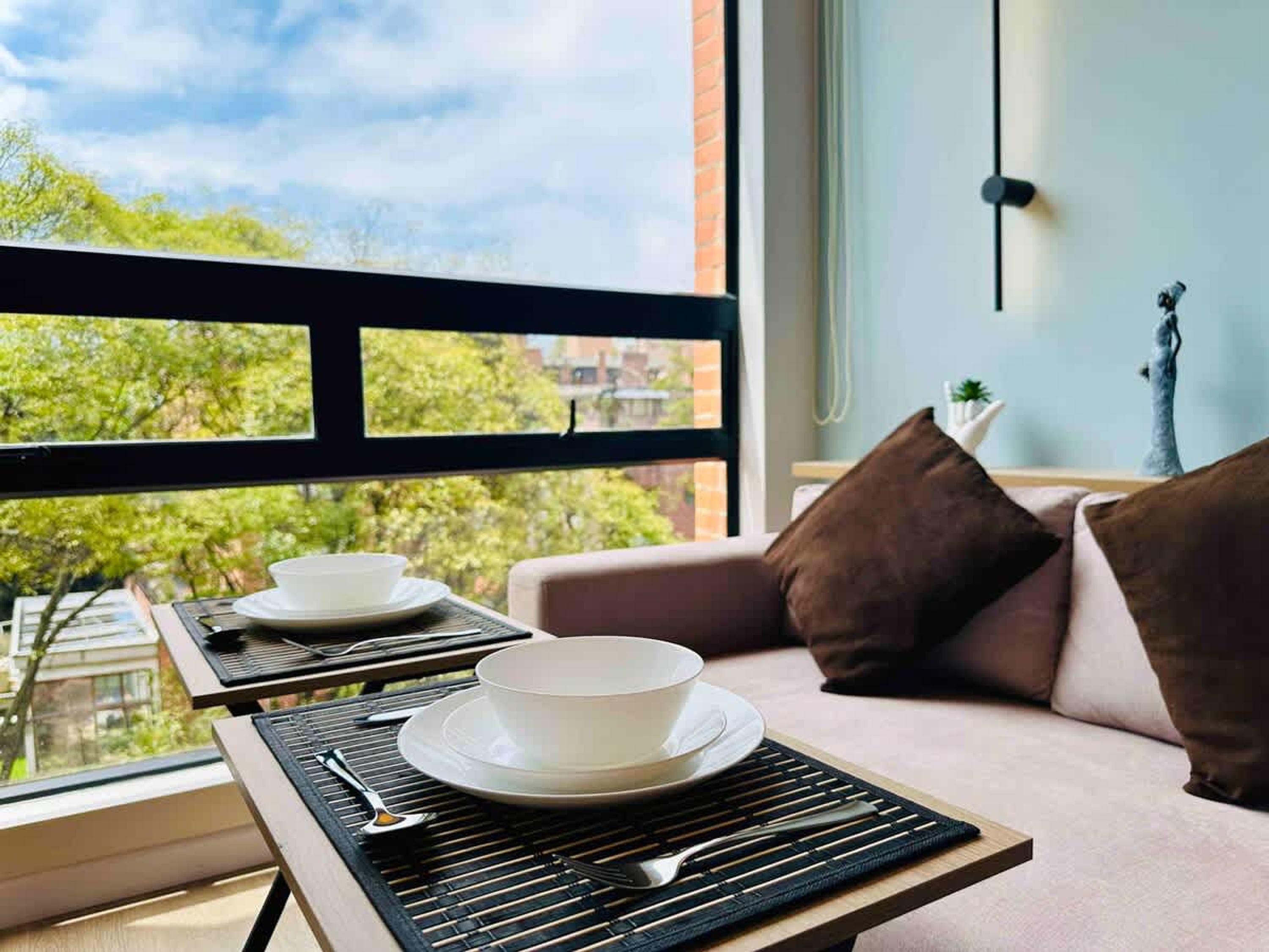Een balkon met een tafel gedekt voor twee personen, een sofa en uitzicht op bomen en gebouwen.