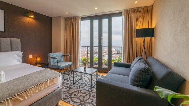 A hotel room with a bed, a sofa, a chair, a small table, and a large window with curtains. - Amazing 1 Br in Candelaria (Bogotá)