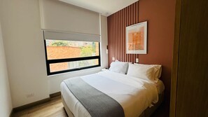 A modern hotel room with a large bed, a window with a view of a brick building, and a framed picture on the wall.