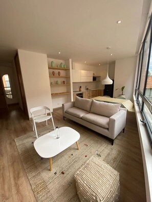 A modern living room with a sofa, a white coffee table, and a woven ottoman. There is a kitchen area with wooden cabinets and a hanging light.