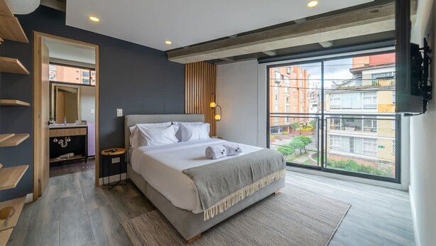 A modern bedroom with a large bed, a bedside table, a mirror, and a window with a view of buildings. - Modern 1 BR in Chapinero (Bogotá)