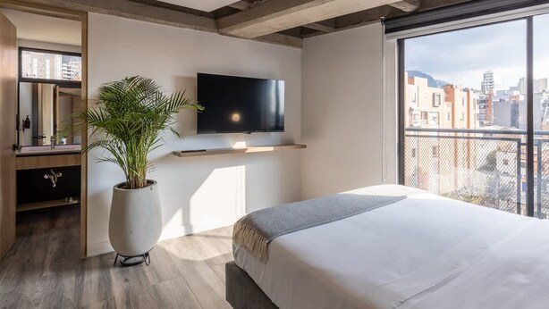 A modern bedroom with a large bed, a TV mounted on the wall, a potted plant, and a balcony with a city view. - Classy 1 BR in Chapinero (Bogotá)