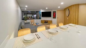 Uma área de jantar moderna com uma mesa branca posta para quatro pessoas, um sofá e uma televisão ao fundo.