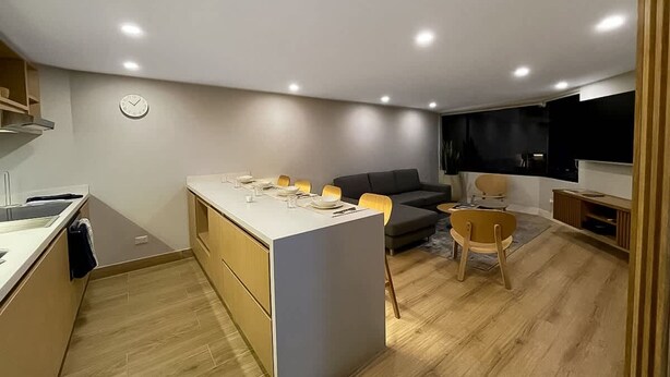Cozinha moderna com ilha central, área de jantar com cadeiras, sofá e televisão na parede.