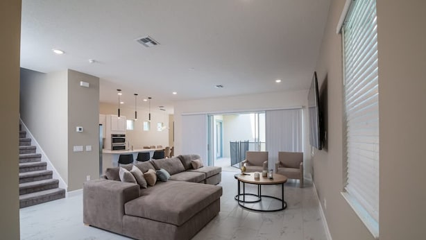 Una sala de estar moderna con un sofá modular, una mesa de centro y un televisor montado en la pared. Se ve una escalera a la izquierda, que conduce a una zona de cocina con armarios y una mesa de comedor.
