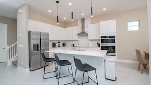 Una cocina moderna con armarios blancos, un refrigerador de acero inoxidable, una isla central con taburetes de bar e iluminación colgante.