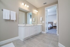 Une salle de bain moderne dotée d’un grand meuble-lavabo, d’une douche à l’italienne et d’une chambre à coucher visible à travers une porte ouverte.