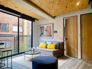 A modern living room with a large window, a sofa with yellow cushions, a round coffee table, and a wooden door. - Panoramic 1 BR in Chapinero (Bogotá)