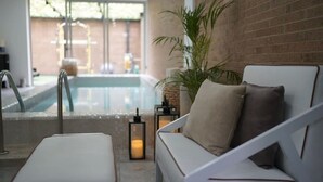 Un espace piscine intérieur moderne, avec un canapé blanc et un fauteuil de relaxation, un mur en briques et une vue sur la piscine.