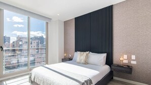 A modern bedroom with a large bed, a city view through floor-to-ceiling windows, a dark headboard, and a nightstand with a lamp.