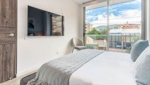 A modern bedroom with a large bed, a TV mounted on the wall, and a view of the city through large windows. - Designer 2BR in Usaqu n (Bogotá)