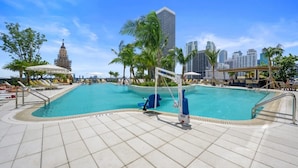 Una piscina con sillas de descanso y sombrillas, rodeada de palmeras y un horizonte urbano de fondo.