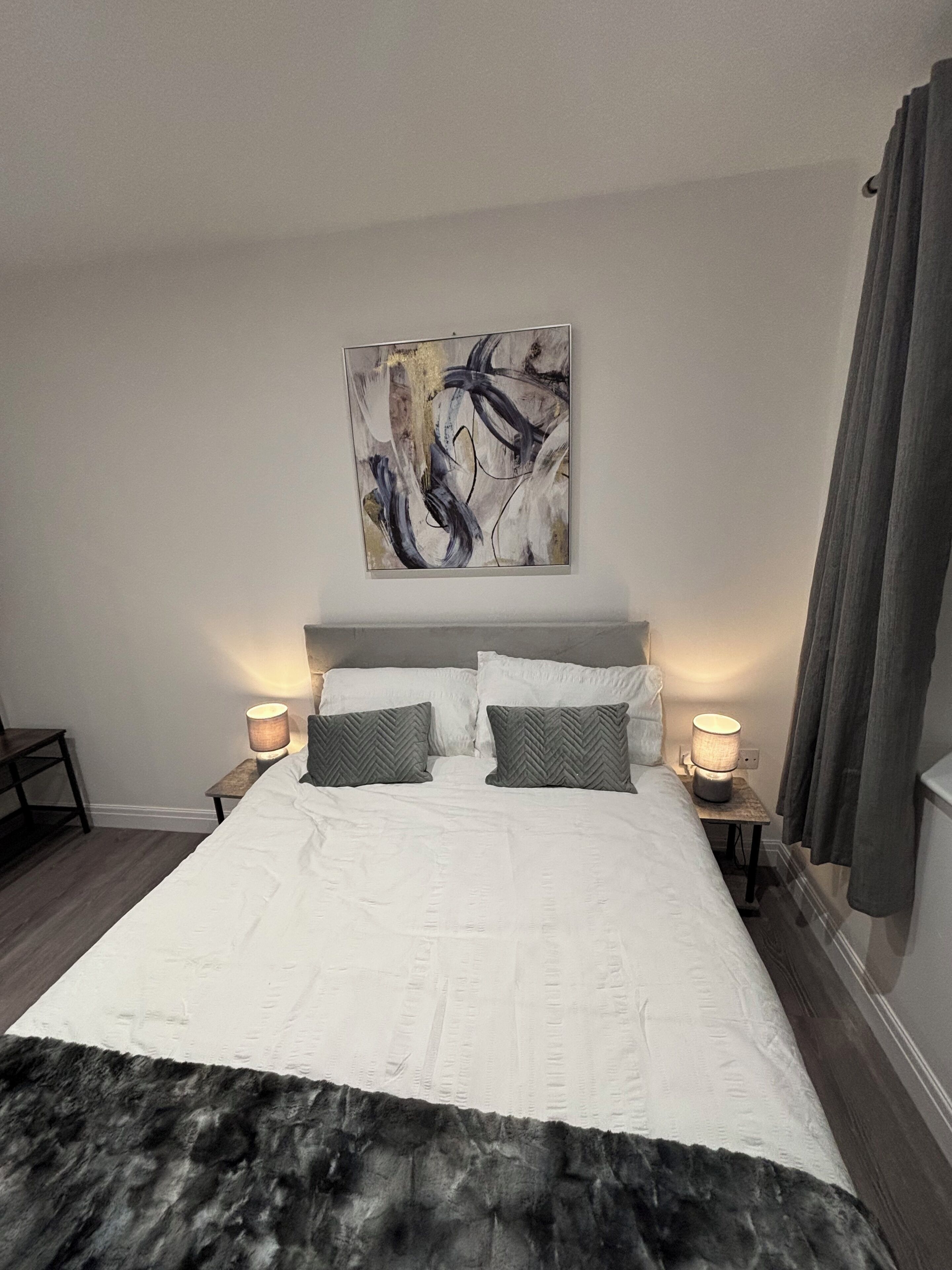 Une chambre avec un grand lit, deux tables de chevet avec des lampes, une peinture abstraite moderne accrochée au mur et un rideau gris.