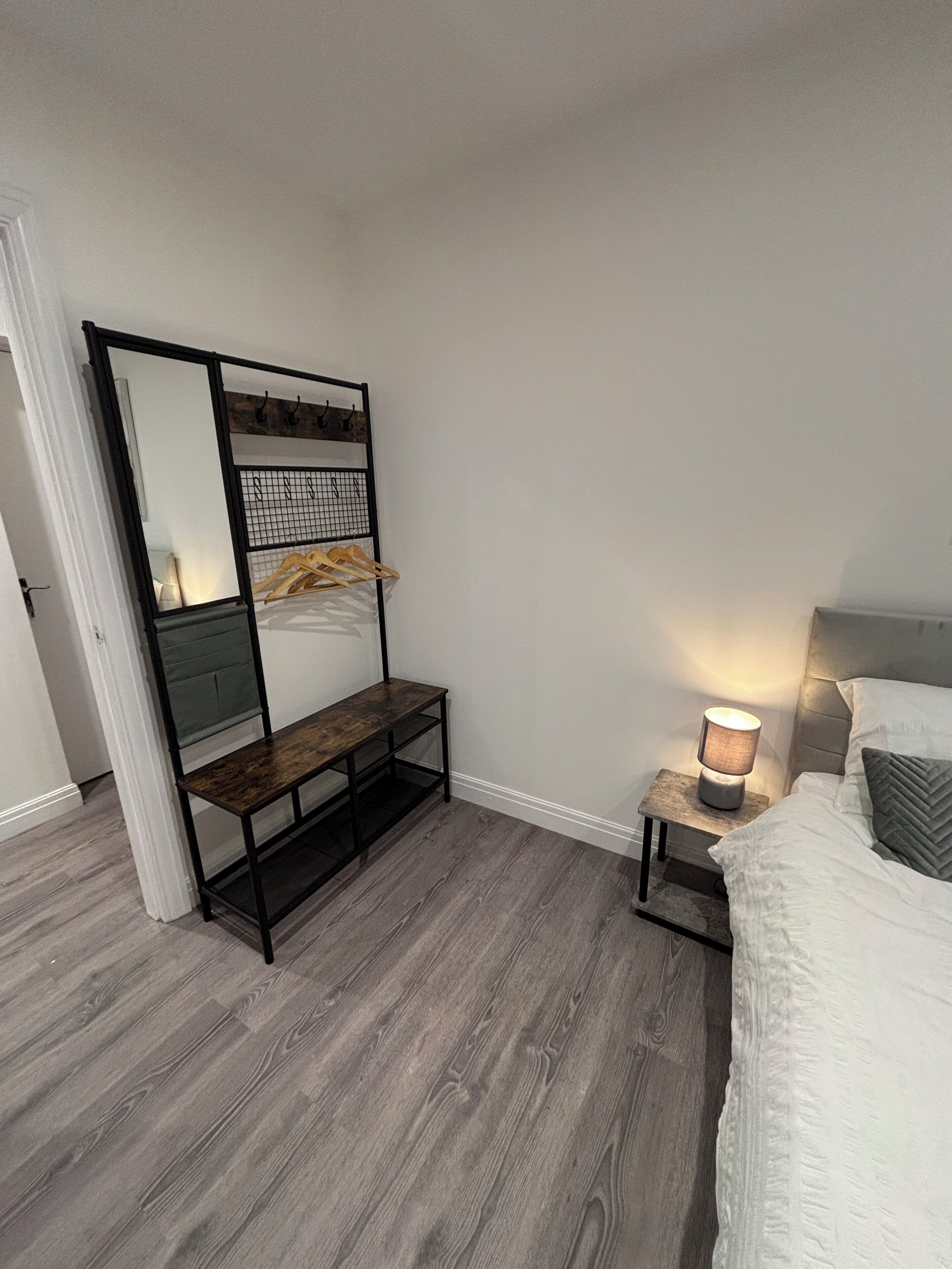 A bedroom with a bed, a bedside table with a lamp, a wooden bench with a mirror, and a coat rack.
