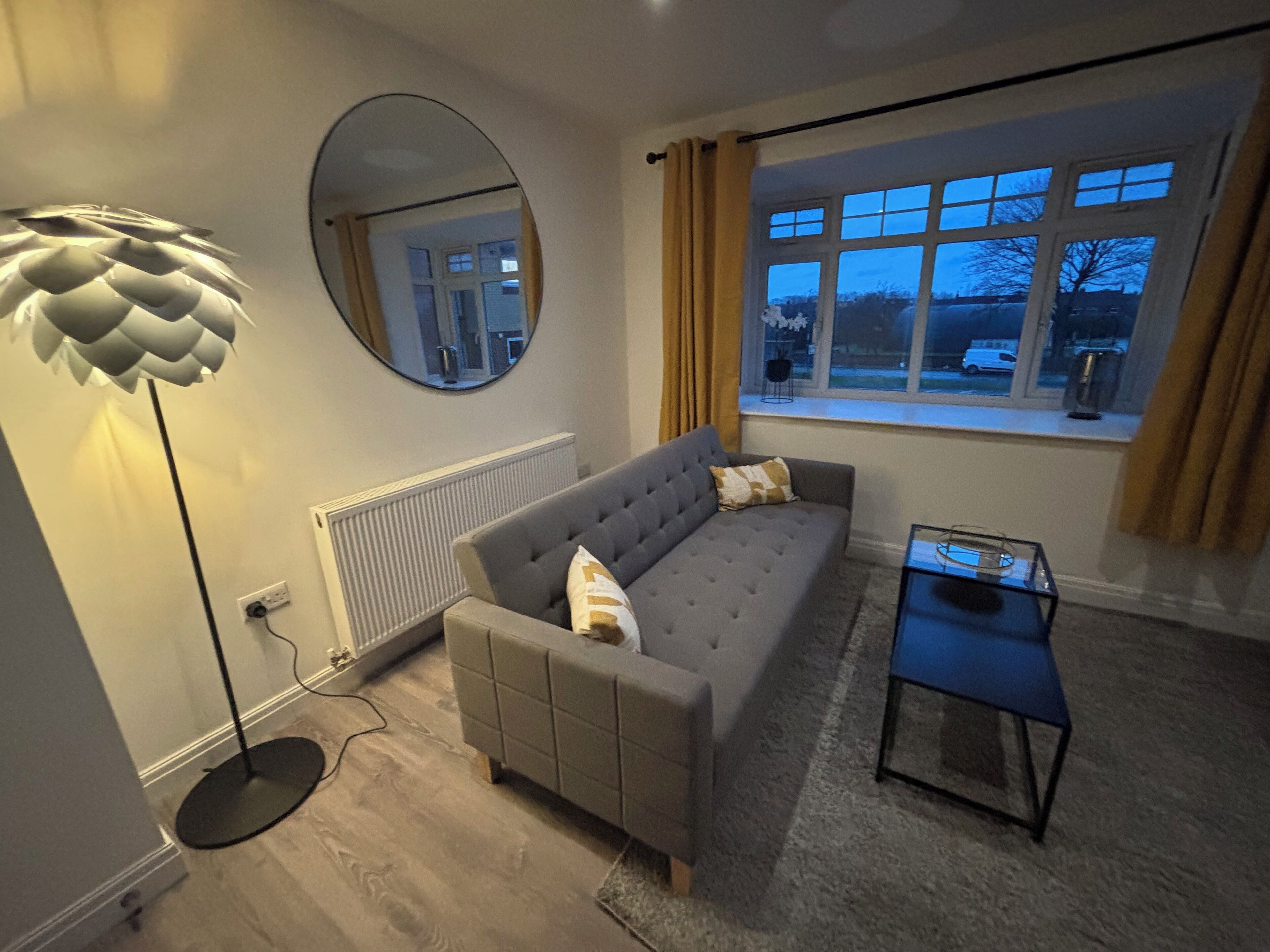 Un salon moderne avec un canapé gris, un miroir rond, une lampe sur pied et une vue sur l’extérieur par une fenêtre.