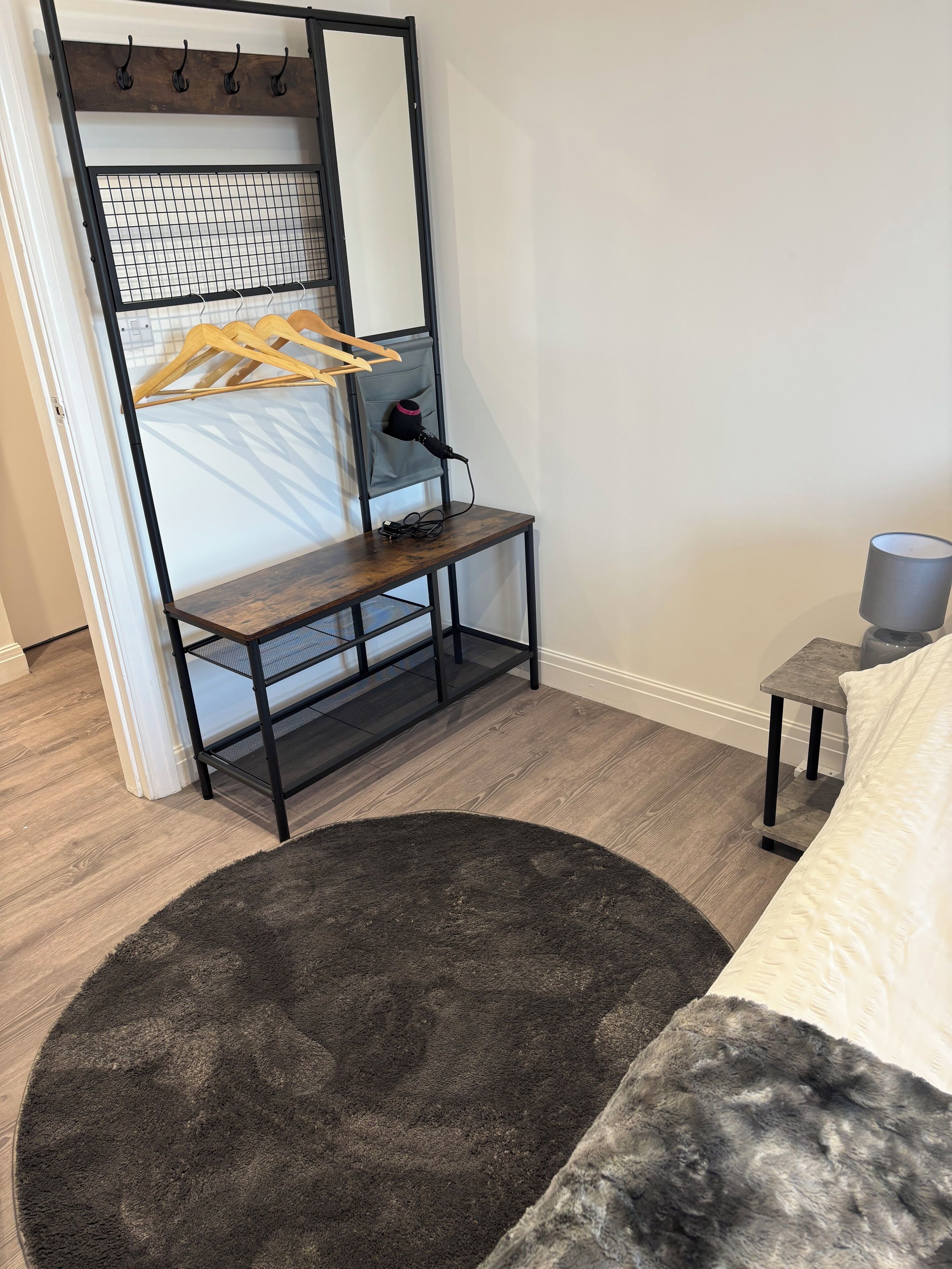 A room with a wooden floor, a dark grey rug, a bed with white bedding, a small table with a lamp, a coat rack with clothes, and a mirror with a grid pattern.