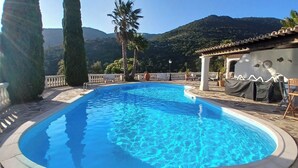 Una piscina con agua azul cristalina, rodeada de baldosas de piedra y una barandilla blanca. Se observan árboles altos y una montaña al fondo.