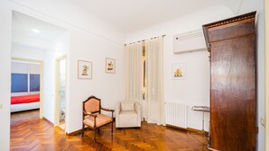 Una habitación con piso de parqué de madera, un armario de madera, una silla, una cama con manta roja y una ventana con vista.