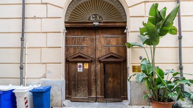 Una puerta doble de madera grande con un arco decorativo en la parte superior, flanqueada por dos botes de basura y una planta en maceta.