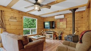 Interior acogedor de una cabaña con paredes de madera, un ventilador de techo, un televisor y un estufa de leña.