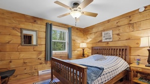 Un dormitorio de cabaña de madera con una cama, una mesita de noche, un ventilador de techo, una ventana con cortinas y un letrero en la pared.