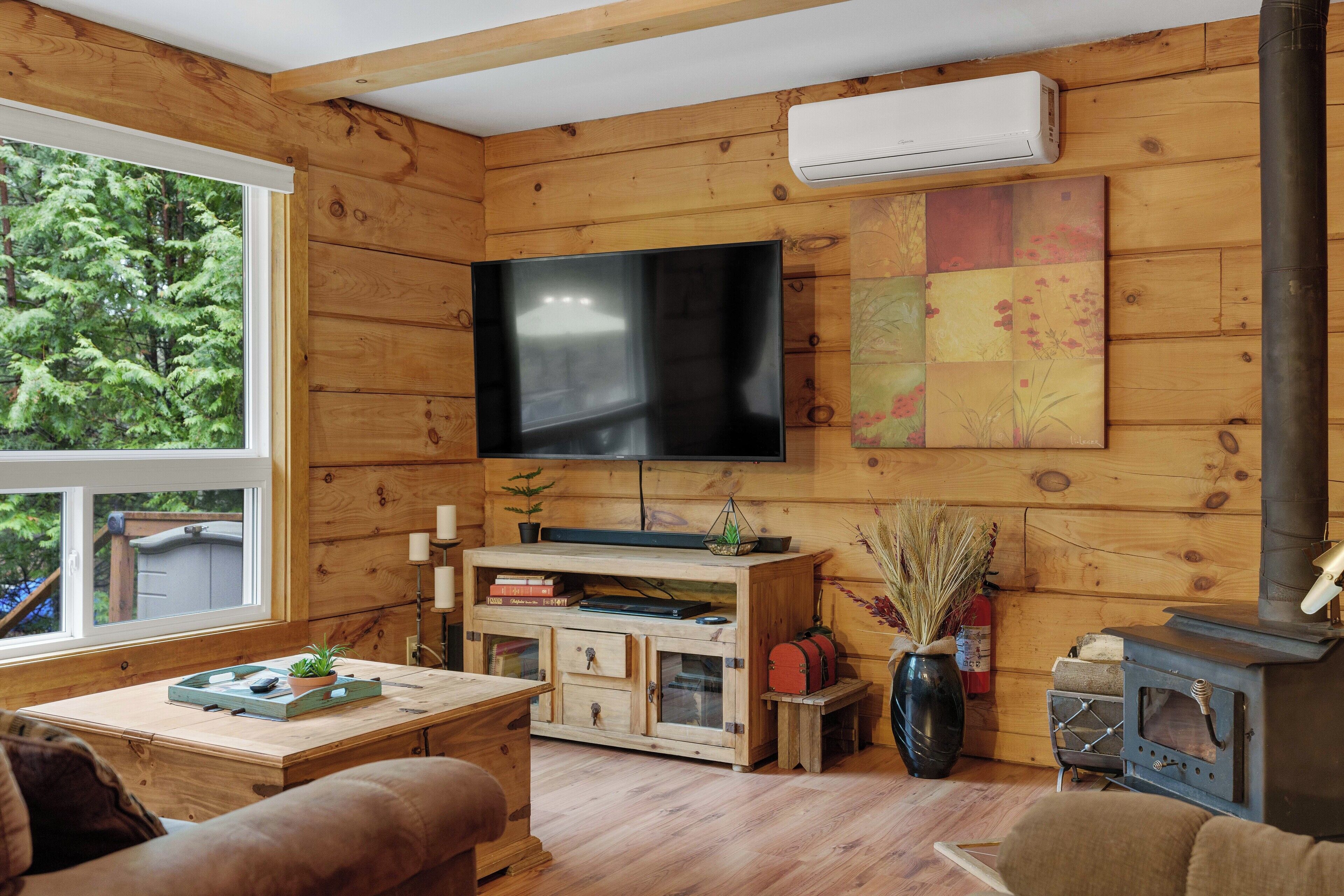 A wooden cabin interior with a TV, wooden furniture, a fireplace, and a window with a view of trees.