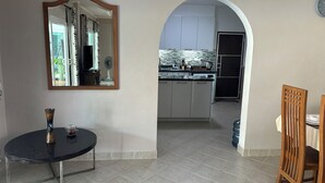 A modern kitchen with white cabinets, a black countertop, and a tiled floor. There is a dining area with a round table and chairs, a mirror on the wall, and a clock on the wall.
