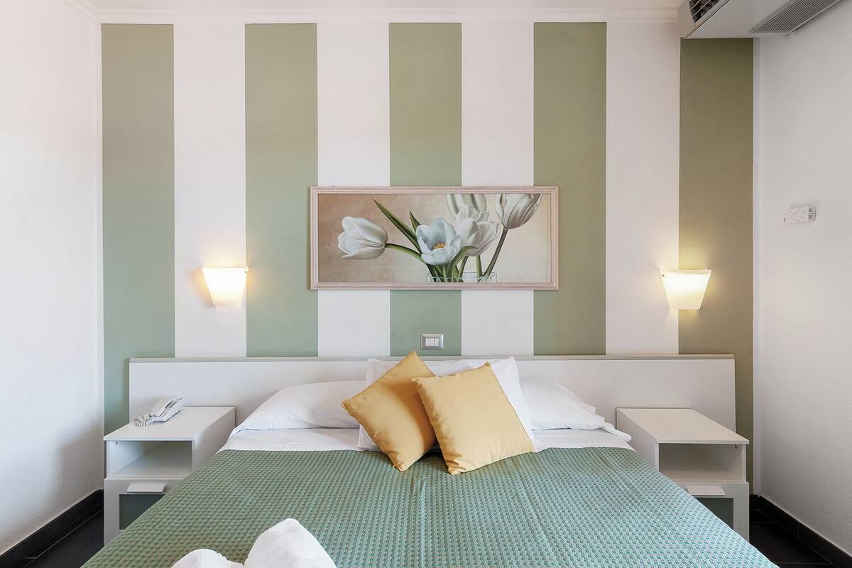 Habitación de hotel con una cama, dos mesitas de noche, una lámpara de noche, una obra de arte floral enmarco y luces fijadas a la pared.