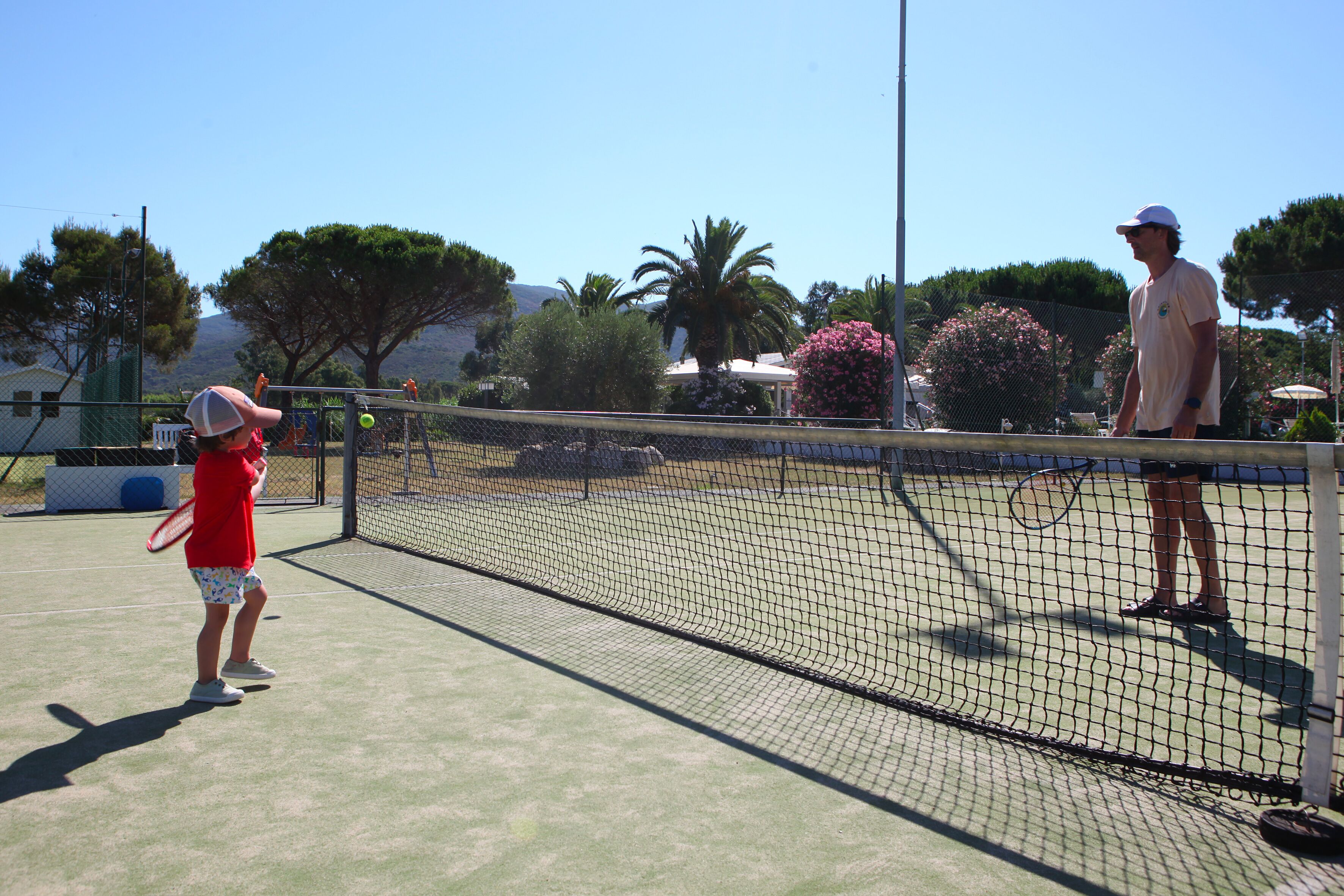 A tennis court with a net, a child in a red shirt, and an adult in a white cap.