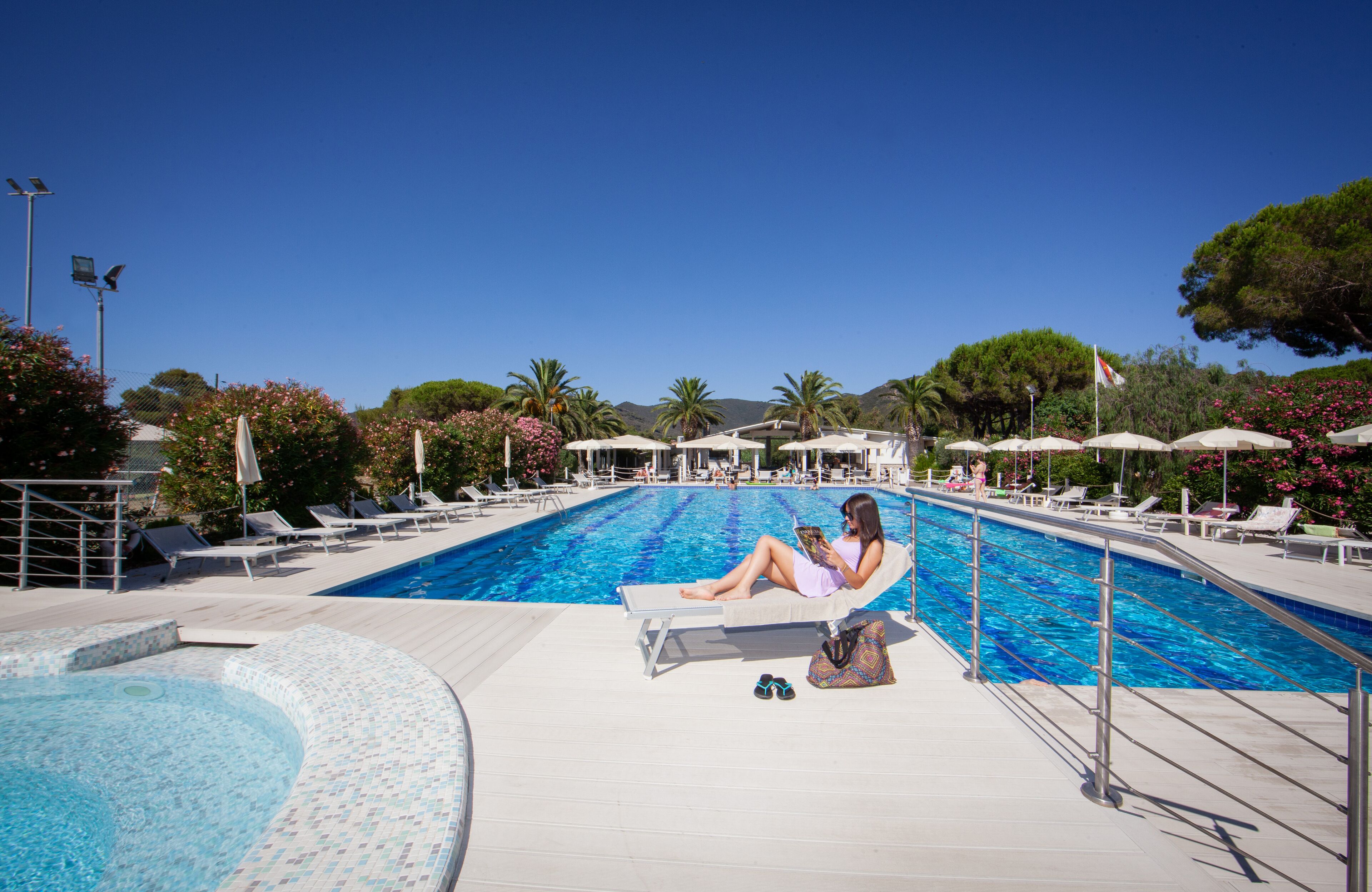 Un espace piscine avec des chaises longues et des parasols, une personne se relaxant sur une chaise, et un ciel bleu dégagé.