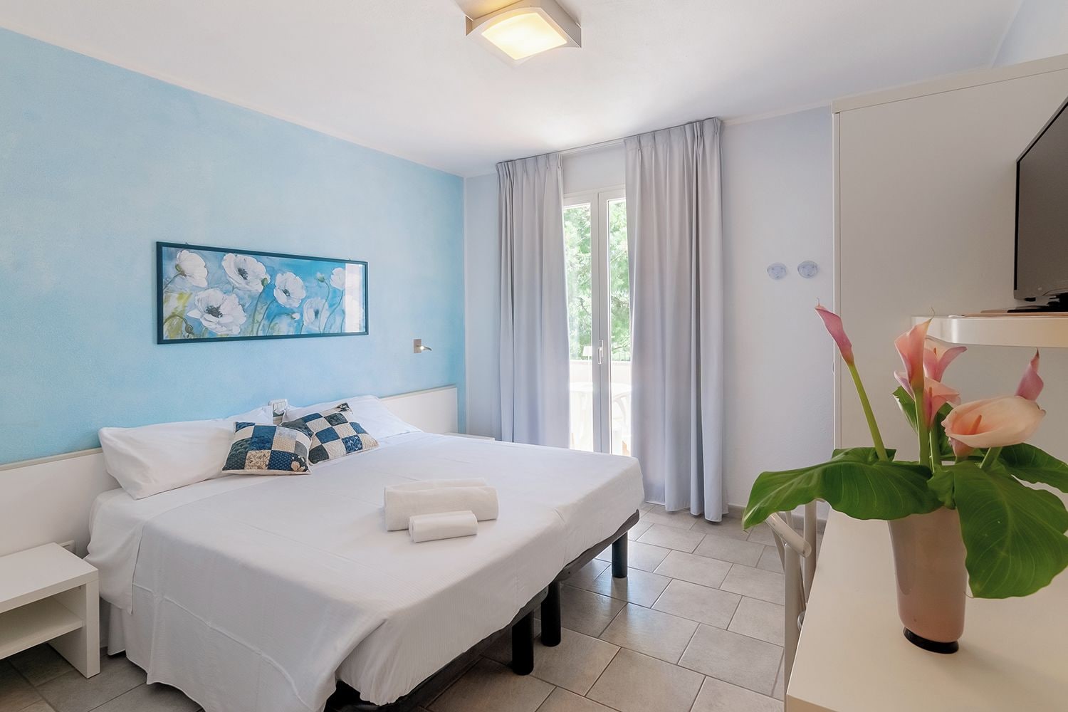 Une chambre d’hôtel avec un lit, une table de chevet, une télévision, une fenêtre avec des rideaux et un tableau au mur.
